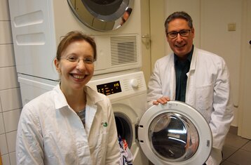 Eine Frau und eine Mann im Laborkittel stehen vor einer offenen Waschmaschine